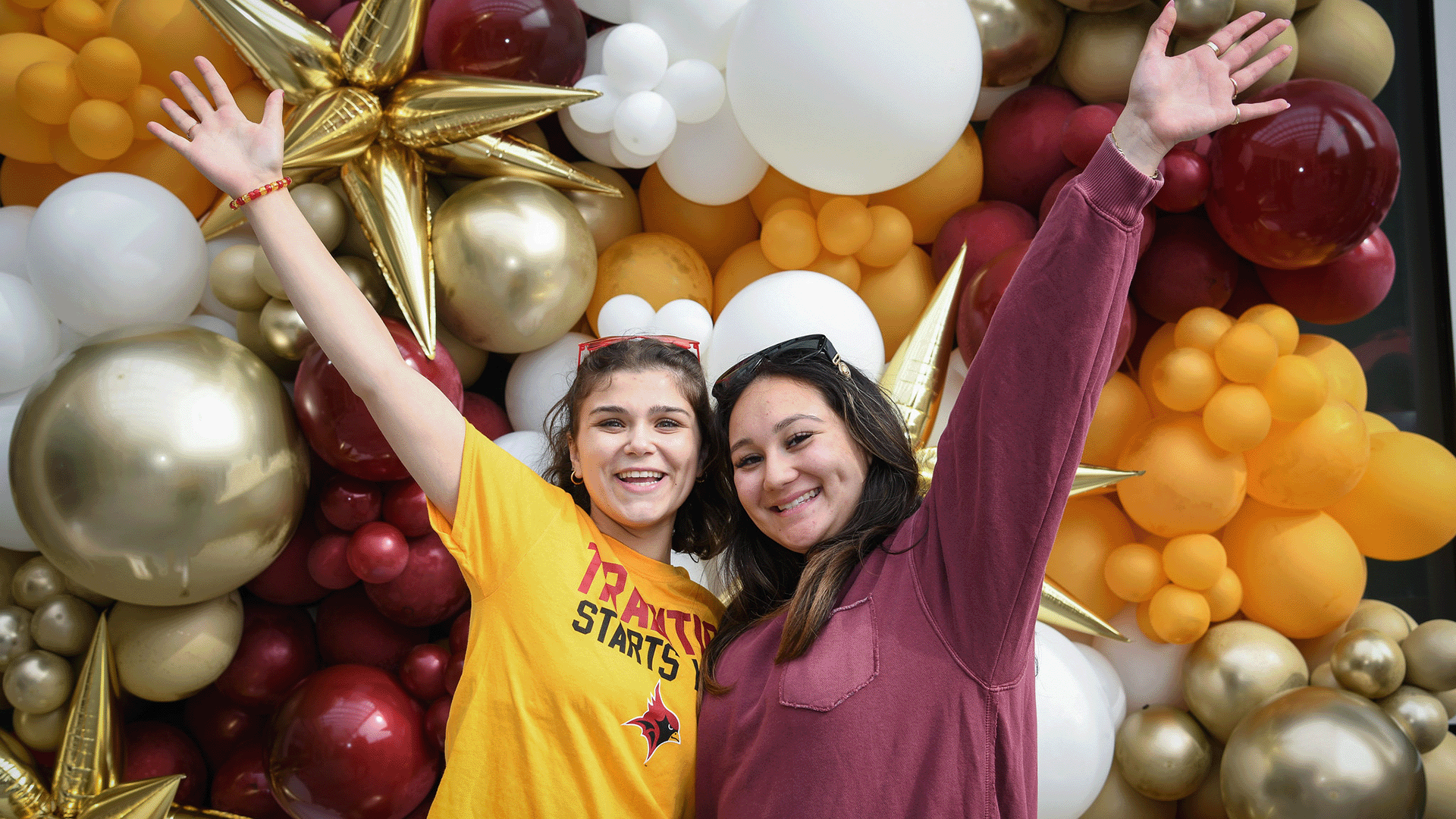 Two students stand with arms raised in excitement in front of wall of balloons.